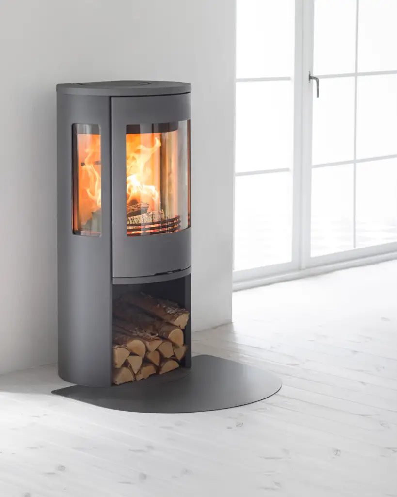 In een Scandinavische woonkamer staat een elegante vrijstaande houtkachel met een gietijzeren deur en grote zijruiten, waarlangs een prachtig vlammenspel zichtbaar is. De kachel, die een tijdloos design heeft, biedt aangename stralingswarmte en voegt een gezellige sfeer toe aan de moderne ruimte.