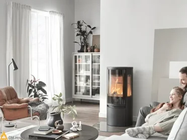 Contura 556 Style ronde, vrijstaande houtkachel met driezijdig vuurzicht en helder oranje vlammen in een Scandinavisch interieur met lichte witte wanden.