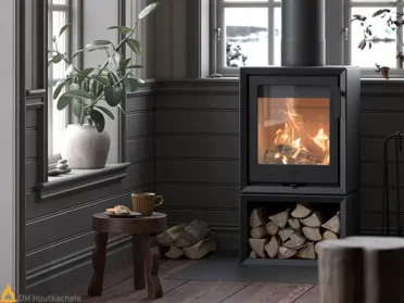 De zwarte Contura 130 Style brandt in een sfeervolle kamer met zicht op het vuur, terwijl onder het toestel een houtvak gevuld is met haardhout.