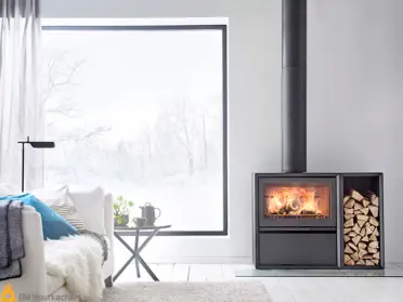 De Contura 310 houtkachel brandt sfeervol naast een houtvak vol gekloofd hout in een lichte Scandinavische woonkamer met groot raam.