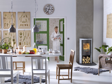 In een lichte woonkamer brandt de Contura 520T Style houtkachel met speksteen en driezijdig vuurzicht naast de eettafel in modern Scandinavisch design.