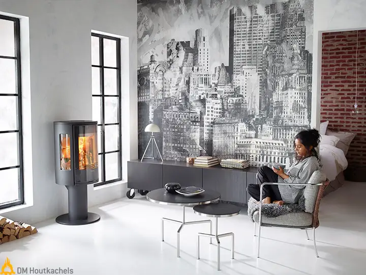 In een moderne woonkamer brandt de zwarte Contura 586G Style houtkachel met driezijdig vuurzicht en glazen deur, naast grote ramen en een zitplek.