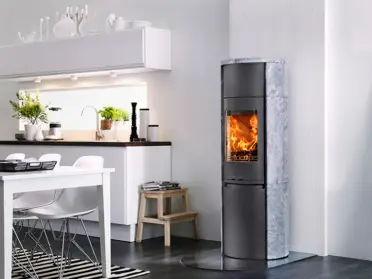 In een moderne eetkamer staat de Contura 590T Style speksteenhoutkachel met ronde vorm en zichtbaar vlammenspel naast de open keuken op donkere vloer.