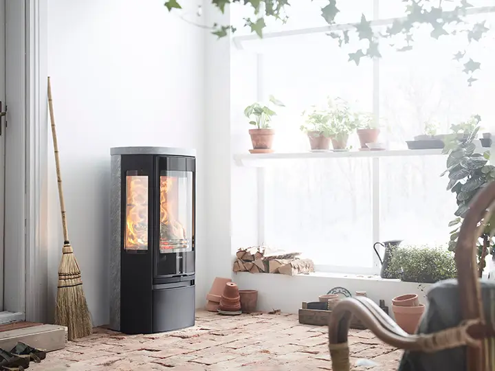 In een lichte serre staat de Contura 856TG Style houtkachel met driezijdig vuurzicht en spekstenen afwerking naast brandhout en kamerplanten.