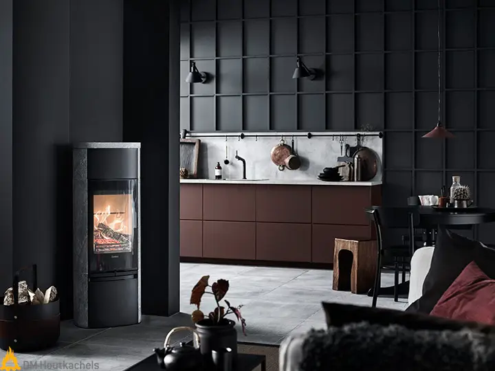 In de donkere woonkamer staat de Contura 890TG Style houtkachel met spekstenen bekleding, terwijl het vuur warm brandt naast een strakke open keuken.