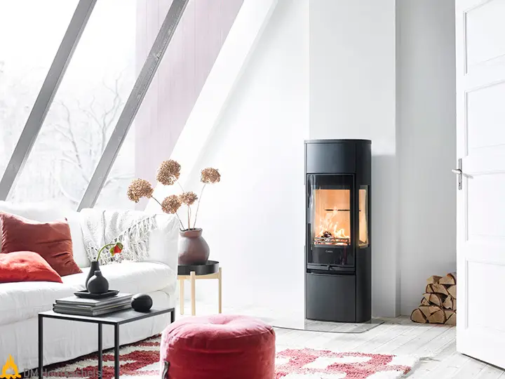 In een lichte woonkamer staat de zwarte Contura 896G Style houtkachel naast een witte wand, terwijl het vuur zichtbaar brandt achter glazen zijruiten.