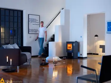 Een man loopt langs een zwarte Contura 52 houtkachel in een modern interieur met houten vloer, zachte verlichting en brandend vuur.