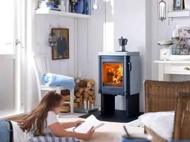 Een meisje leest op de vloer bij de Contura 52T houtkachel, waar het vuur warm brandt in een lichte woonkamer met witte muren en houten blokken.