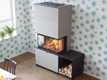 Een Contura i51AN houtkachel brandt in een modern interieur met zwart-witte ombouw, houtopslag en turquoise bloemenbehang naast een raam.
