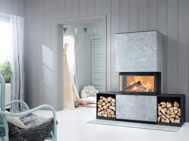 De Contura i51T houtkachel met speksteen brandt sfeervol naast opgestapeld brandhout in een lichte woonkamer met grijze wanden en brede ramen.